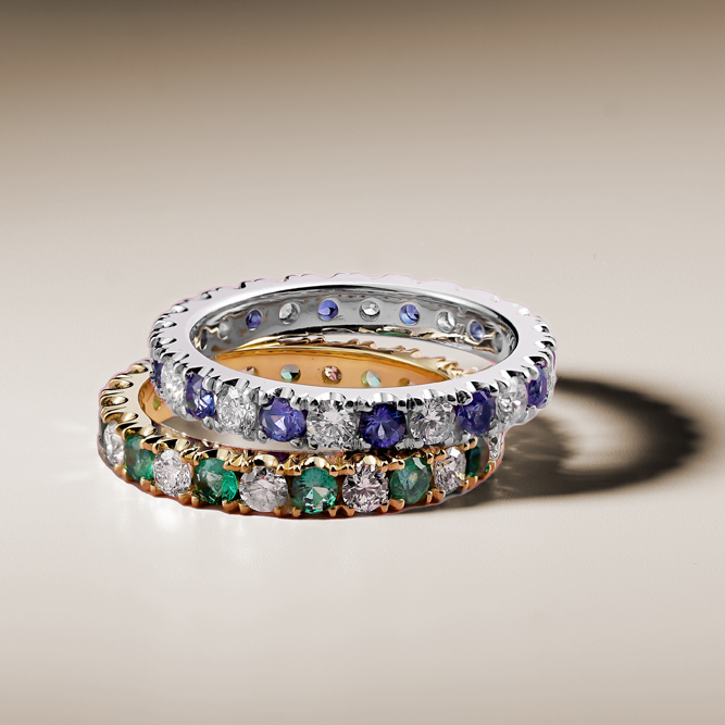 Two rings stacked on top of each other. On the bottom, the 'Always' ring, a 18K yellow gold ring featuring a seamless blend of 0.65tcw round cut emeralds and 0.72tcw diamonds in prong settings. On top, a similar ring, in silver.