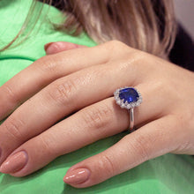 Load image into Gallery viewer, Stunning engagement ring in 18K white gold, featuring a vibrant cushion-cut Ceylon sapphire, surrounded by 14 round brilliant diamonds totaling 0.70tcw, with double eagle claw settings, handcrafted in Montreal by Exaurum.
