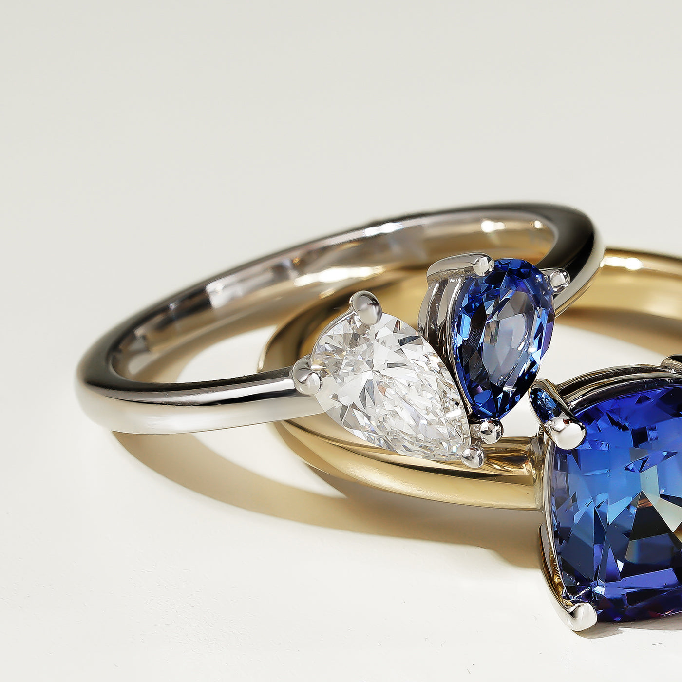 Two rings stacked on top of each other. The top, an elegant two stone ring in 14K white gold, featuring a 0.56ct Chatham sapphire and a 0.45ct lab diamond pear shape, VS/F, in a 3-prong setting, handcrafted in Montreal by Ex Aurum. Below it, another sapphire ring set in yellow gold.